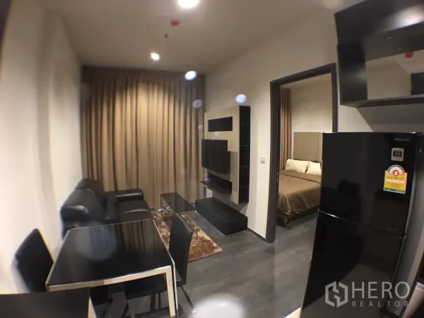 คอนโด ให้เช่าในWatthana, Bangkok - ห้องนั่งเล่นขนาดกะทัดรัด โซฟาหนังสีดำ ชุดโต๊ะอาหาร ผนังทีวี และประตูสู่ห้องนอน พร้อมตู้เย็นขนาดใหญ่