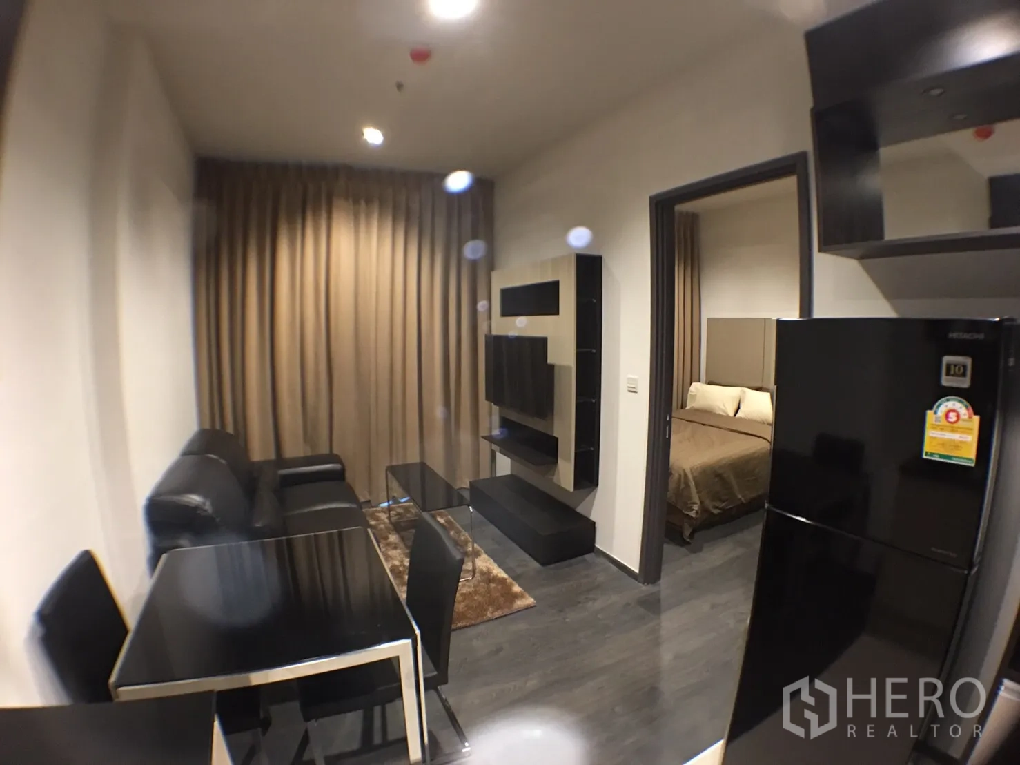 คอนโด ให้เช่าในWatthana, Bangkok - ห้องนั่งเล่นขนาดกะทัดรัด โซฟาหนังสีดำ ชุดโต๊ะอาหาร ผนังทีวี และประตูสู่ห้องนอน พร้อมตู้เย็นขนาดใหญ่