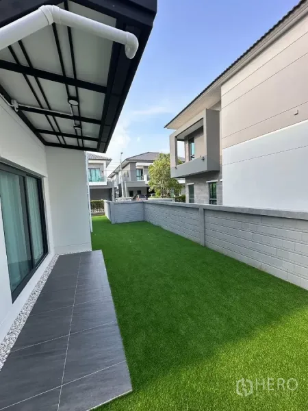 Detached House for rent in Bang Phli, Samut Prakan - Side garden with artificial turf, tiled terrace, boundary wall, and roof overhang.