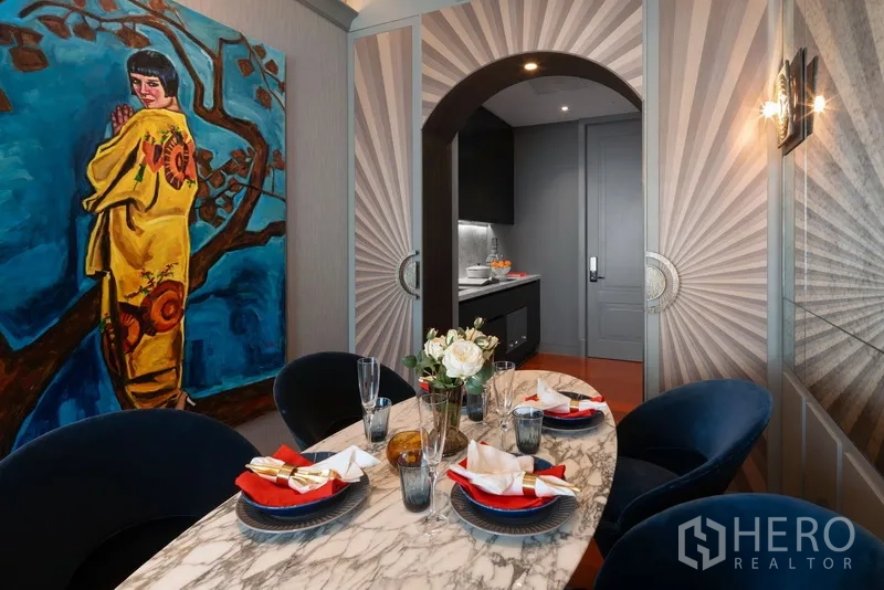 Condominium for sale in Watthana, Bangkok - Marble‑top dining table set for four beside bold wall art and an arched doorway to the kitchen.