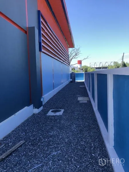 货栈 出租或出售在Sai Noi, Nonthaburi - 仓库外侧碎石步道，沿蓝色围墙延伸。