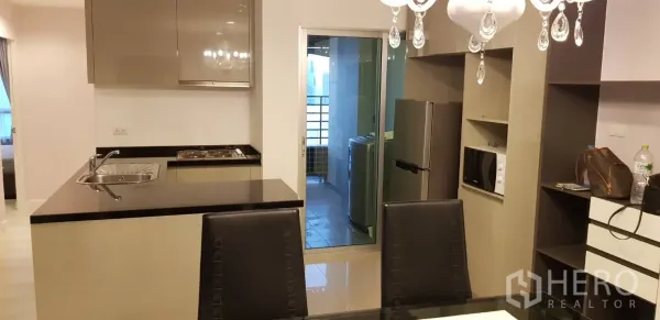 Condominium for rent in Huai Khwang, Bangkok - Modern kitchen with black countertop island, chandelier, and a sliding glass door to a laundry balcony.