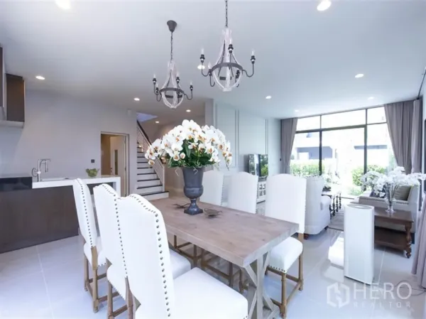 Detached House for rent in Bang Kapi, Bangkok - Dining space with wooden table, white upholstered chairs, and pendant lights beside the kitchen and stairs.