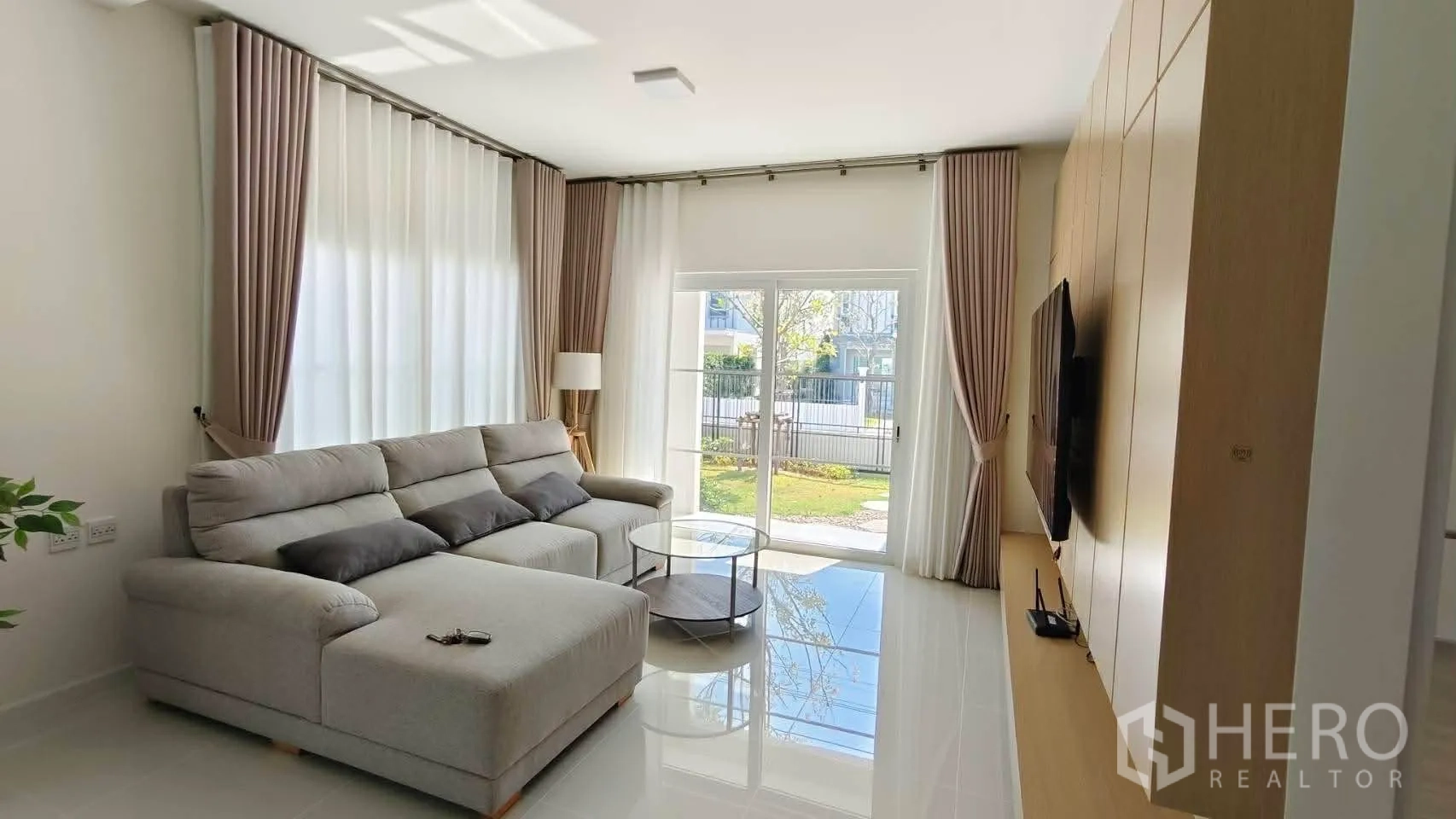 独立式住宅 for rent in Bang Phli, Samut Prakan - 采光充足的客厅，L形沙发，落地推拉门通向花园，内置电视墙。