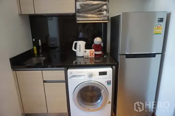Condominium for rent or sale in Khlong Toei, Bangkok - Compact kitchenette with black countertop, washing machine, microwave and stainless fridge in the unit.