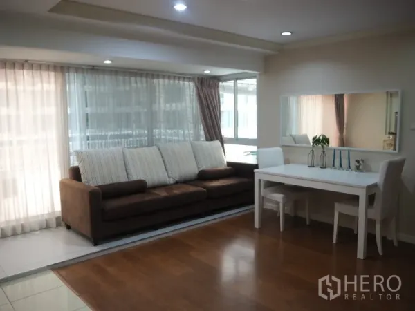 Condominium for rent or sale in Watthana, Bangkok - Lounge with large brown sofa, floor-to-ceiling windows with sheer curtains, and a dining nook.
