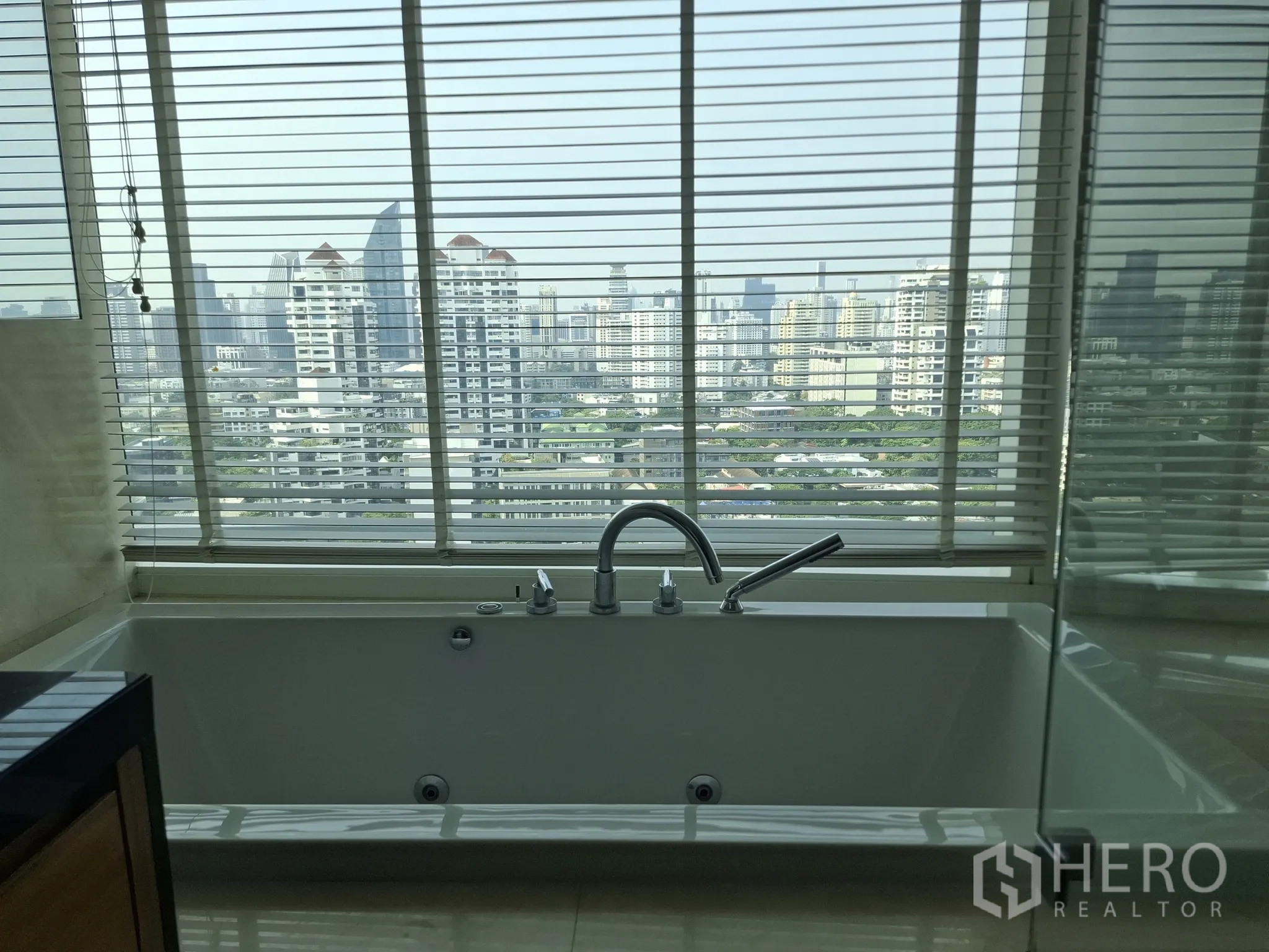 Condominium for rent in Watthana, Bangkok - Bathtub by window with panoramic Bangkok city view through blinds.