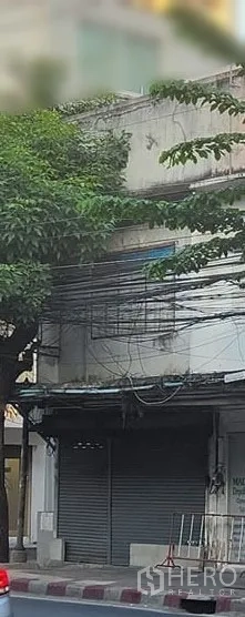 店屋 出租在Watthana, Bangkok - 两层街铺外立面，金属卷闸门紧闭，临街人行道与绿树，位于曼谷Ekkamai城区的城市景观。