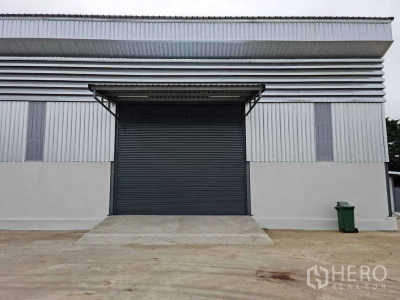 货栈 出租在Bang Bua Thong, Nonthaburi - 仓库正立面，金属板外墙，超大卷闸门与带雨棚的装卸区，前方为宽阔水泥场地。
