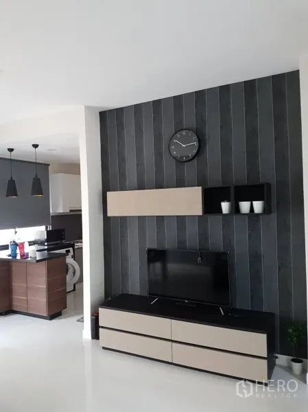 Townhouse for rent or sale in Saphan Sung, Bangkok - Living room with TV console, wall clock and view into the kitchen in a bright townhouse.