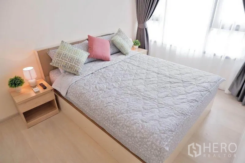 Condominium for rent in Huai Khwang, Bangkok - Sunlit master bedroom with large windows, gray bedding, and full curtains.