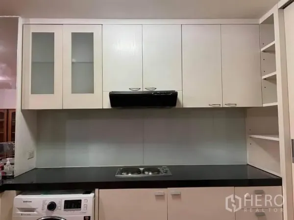 Condominium for rent or sale in Watthana, Bangkok - Modern white kitchen with glass-front cabinets, electric cooktop, range hood, and an in-unit washing machine.