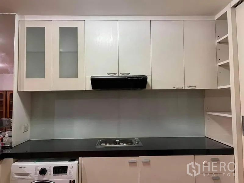 Condominium for rent or sale in Watthana, Bangkok - Modern white kitchen with glass-front cabinets, electric cooktop, range hood, and an in-unit washing machine.