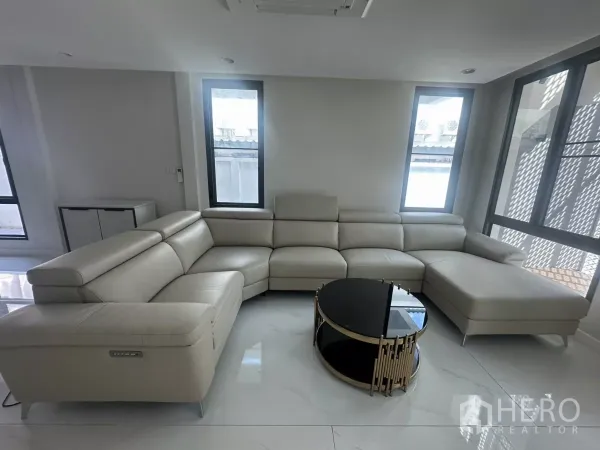 联排别墅 出租在Watthana, Bangkok - 明亮客厅，米色L形真皮沙发与圆形茶几，落地窗采光。