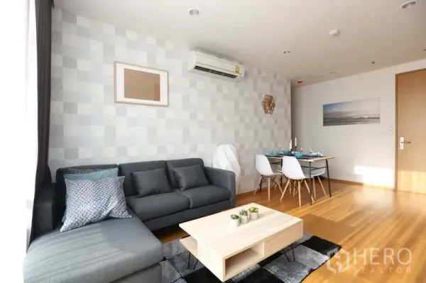 Condominium for sale in Bang Rak, Bangkok - Cozy living room with gray sectional sofa, wall-mounted air conditioner and dining nook.