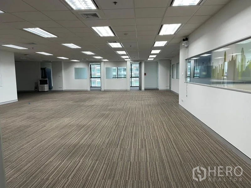Office Space for rent - Bright open workspace with fluorescent lights, glass partitions and windows at the far end.