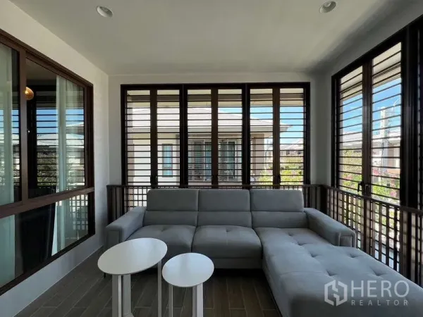Detached House for rent in Bang Kapi, Bangkok - Enclosed balcony lounge with gray sectional sofa and louvered windows.