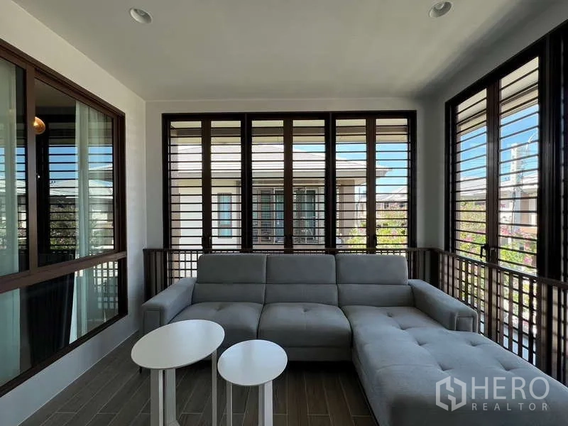 Detached House for rent in Bang Kapi, Bangkok - Enclosed balcony lounge with gray sectional sofa and louvered windows.