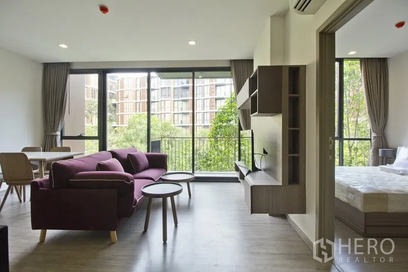 คอนโด ให้เช่าในWatthana, Bangkok - โถงพักผ่อนแบบเปิดโล่งมีโซฟาสีม่วง ระเบียงวิวต้นไม้ ประตูเลื่อนไปยังห้องนอน และชุดรับประทานอาหารริมหน้าต่างสูง