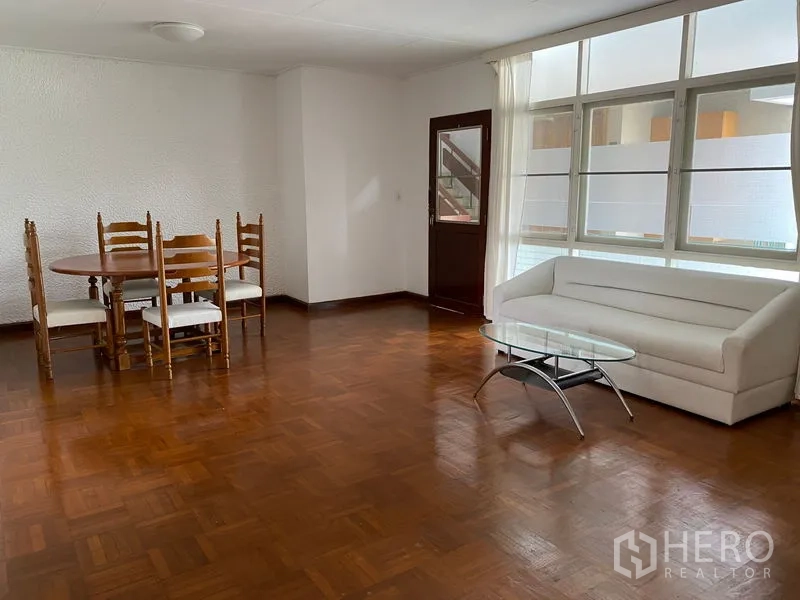 Apartment for rent in Khlong Toei, Bangkok - Spacious living and dining area with parquet floors, white sofa, glass coffee table and large windows.