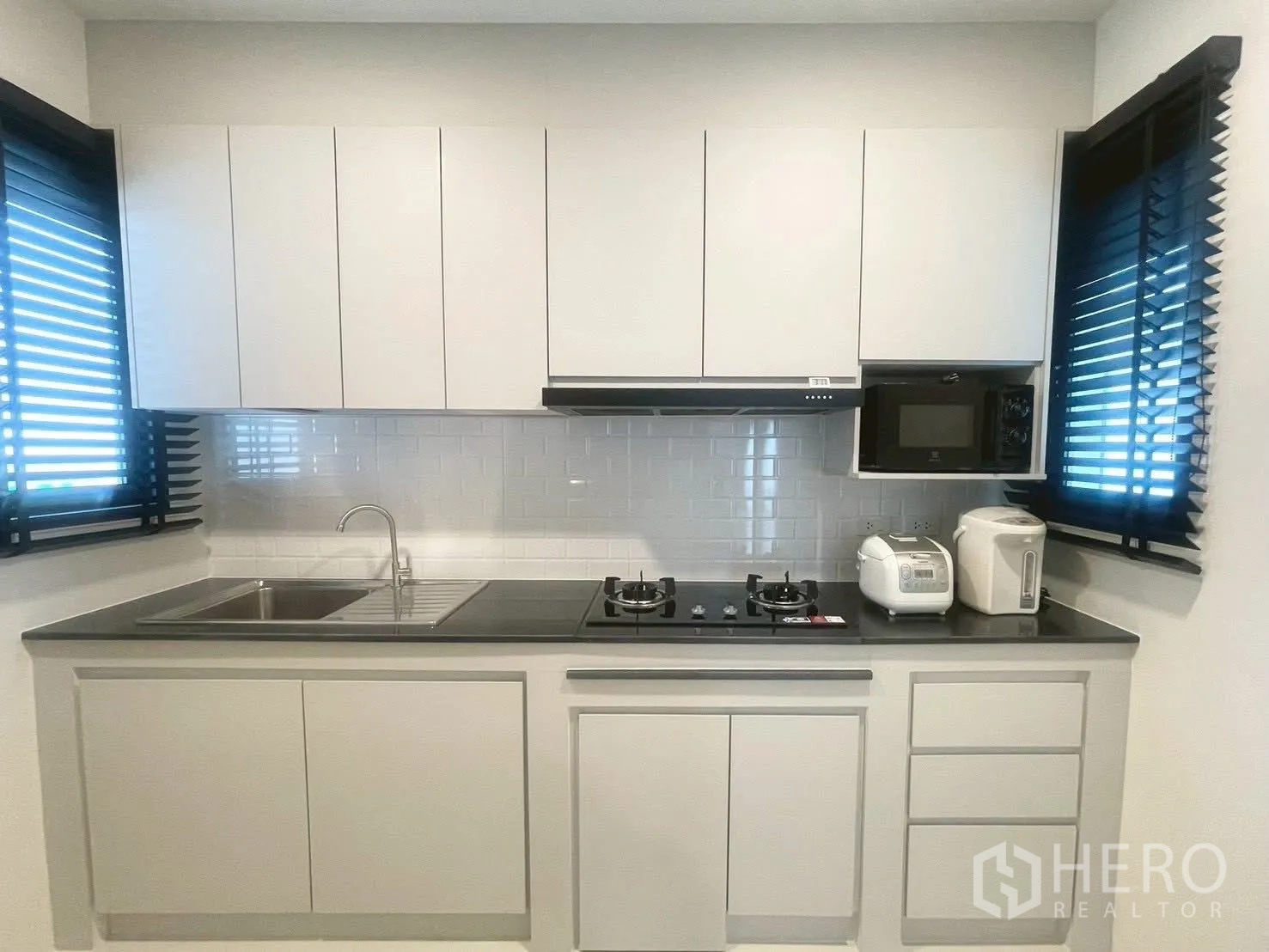 Detached House for rent in Bang Phli, Samut Prakan - Modern white kitchen with gas hob, sink, microwave and windows with blinds.