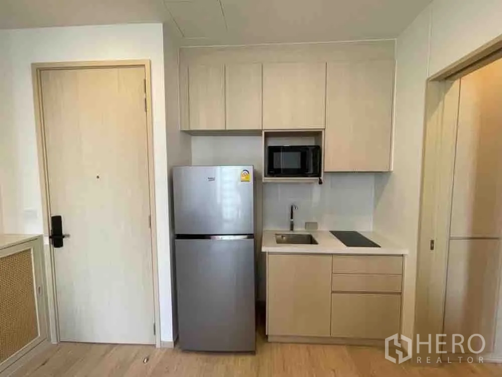 公寓 for rent in Watthana, Bangkok - 紧凑厨房配浅色橱柜、水槽、电磁炉、微波炉及不锈钢冰箱，旁为入户门。