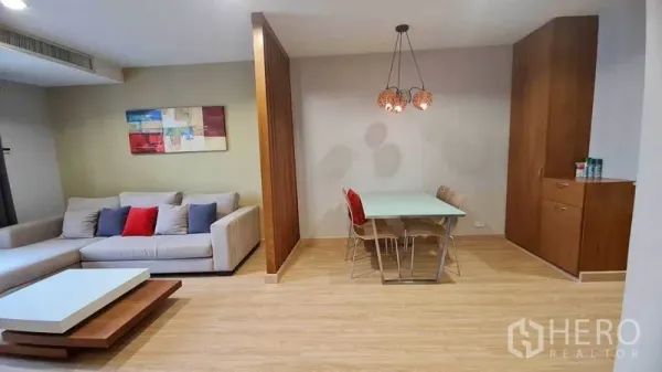 Condominium for rent or sale in Watthana, Bangkok - Modern lounge with L-shaped sofa, wall art, and a dining nook divided by a wooden screen.