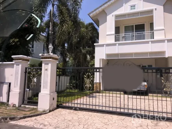 บ้าน for ในLat Krabang, Bangkok - บ้านเดี่ยวสองชั้นสีขาวในย่านลาดกระบัง กรุงเทพฯ มีระเบียง ประตูรั้วเหล็ก ถนนปูพื้น ที่จอดรถในร่ม และสนามหญ้าพร้อมต้นปาล์ม.