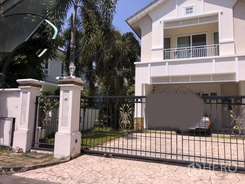 บ้าน for ในLat Krabang, Bangkok - บ้านเดี่ยวสองชั้นสีขาวในย่านลาดกระบัง กรุงเทพฯ มีระเบียง ประตูรั้วเหล็ก ถนนปูพื้น ที่จอดรถในร่ม และสนามหญ้าพร้อมต้นปาล์ม.