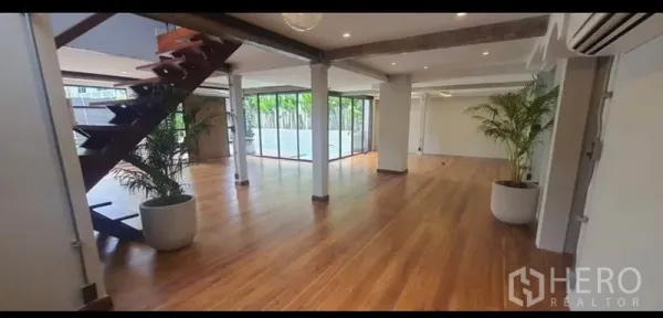 Home Office for rent or sale in Khlong Toei, Bangkok - Spacious open-plan interior with wooden floors, exposed beams, a staircase and floor-to-ceiling windows facing greenery.