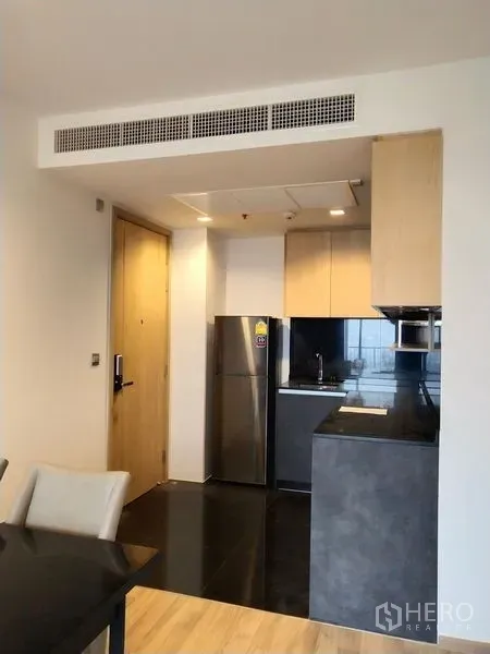 Condominium for rent in Chatuchak, Bangkok - Open-plan kitchen with light wood cabinets, black countertop, and a stainless refrigerator beside the entry.