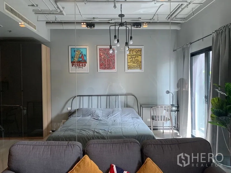 Condominium for sale in Watthana, Bangkok - View of the bedroom through a glass partition from the sofa, with artwork above the bed and large windows.
