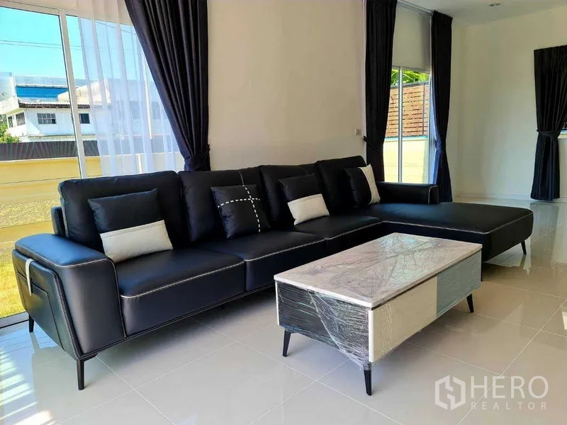 Detached House for rent or sale in Pathum Thani - Sunlit living room with black sectional sofa beside tall sliding windows and sheer curtains.