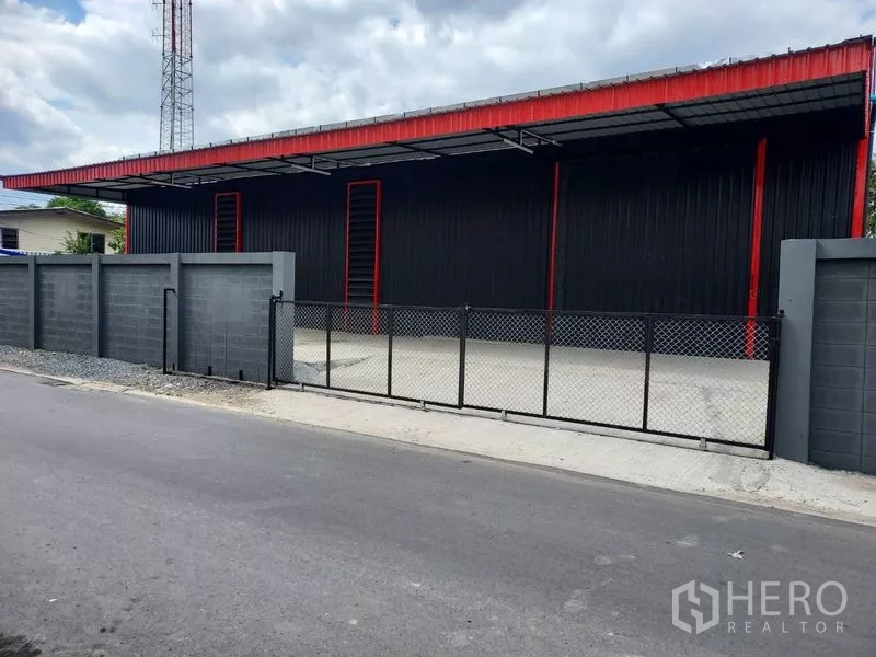 คลังสินค้า ให้เช่าในBang Kapi, Bangkok - มุมมองจากถนนของโกดังสีดำ–แดง พร้อมรั้วเหล็กและลานโหลดของมีหลังคา