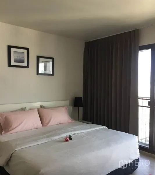 Condominium for rent in Watthana, Bangkok - Cozy bedroom with king bed, pink pillows, bedside table and lamp, blackout curtains, and balcony access.