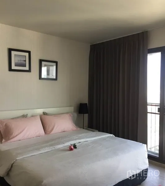 Condominium for rent in Watthana, Bangkok - Cozy bedroom with king bed, pink pillows, bedside table and lamp, blackout curtains, and balcony access.