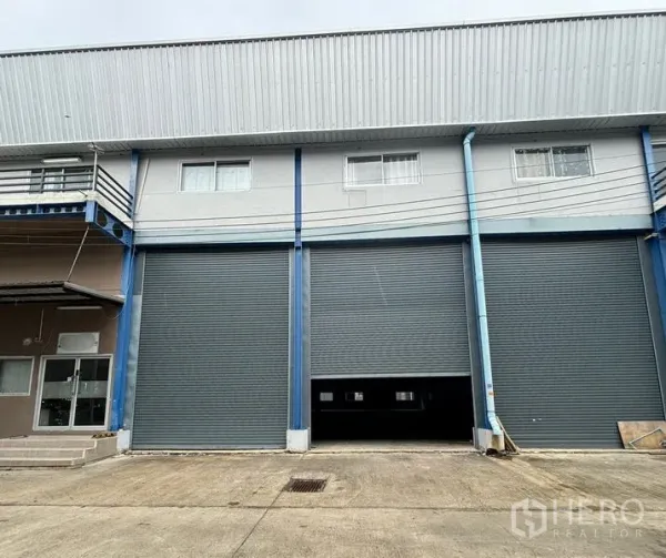 Warehouse for rent in Krathum Baen, Samut Sakhon - Street-level view of the warehouse exterior with closed roller shutters and parking area.