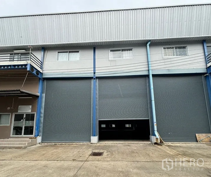 Warehouse for rent in Krathum Baen, Samut Sakhon - Street-level view of the warehouse exterior with closed roller shutters and parking area.