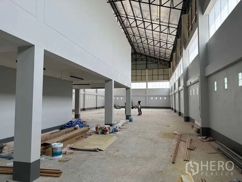 Factory for sale in Khlong Luang, Pathum Thani - Wide factory bay with interior columns, tall windows and construction materials on the floor.