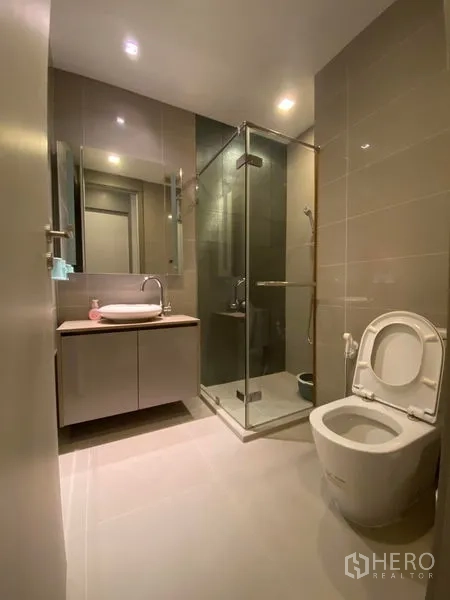 Condominium for rent in Bang Rak, Bangkok - Modern bathroom with glass shower, vanity, and toilet in neutral tiles.