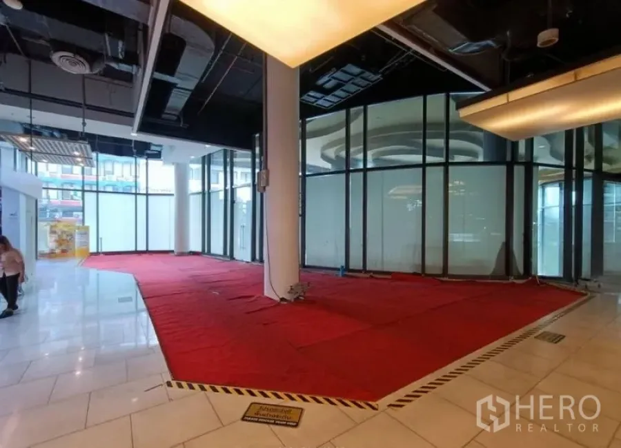 Retail Space for rent in Khlong Toei, Bangkok - Vacant retail corner with wraparound windows, high ceiling lighting and red mat flooring near a mall entrance.