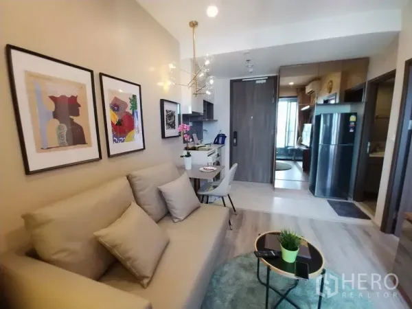 Condominium for rent in Huai Khwang, Bangkok - Open-plan living area facing the entry with sofa, round rug, compact kitchen, and mirrored storage cabinets.