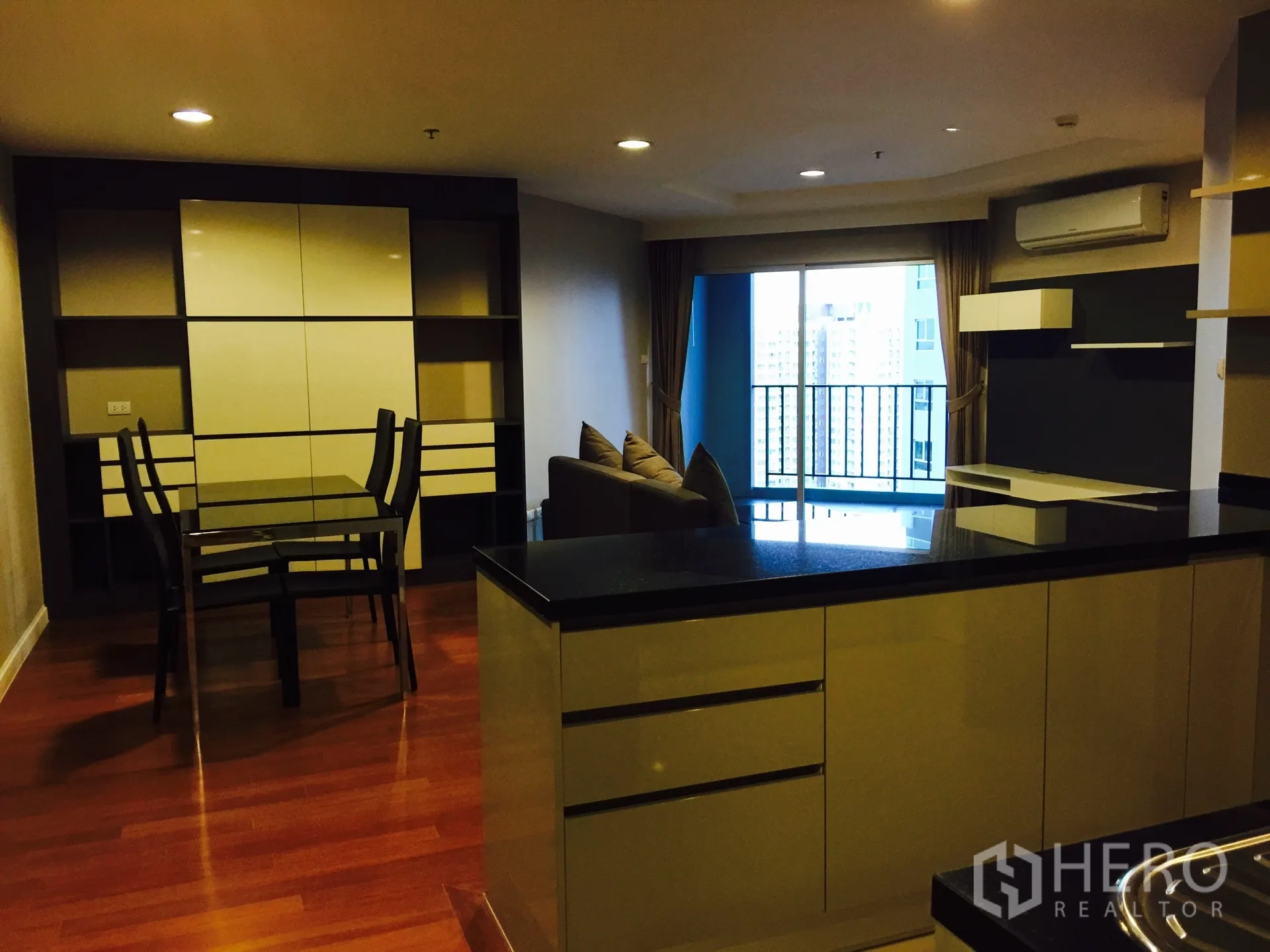Condominium for rent in Huai Khwang, Bangkok - Open-plan kitchen with black countertops, built-in cabinets, dining table and view to the balcony.