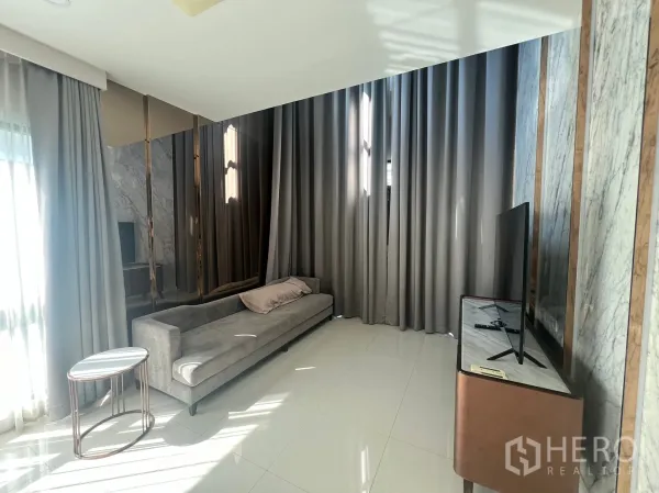 บ้าน ให้เช่าหรือขายในPrawet, Bangkok - ห้องนั่งเล่นสว่าง โซฟายาวสีเทา ผ้าม่านสูงจรดเพดาน และชั้นวางทีวี