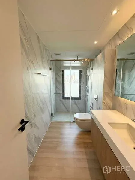 Condominium for rent in Watthana, Bangkok - Modern bathroom with marble walls, long vanity, glass-enclosed rain shower, and city-view window.