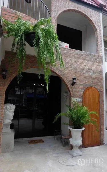 联排别墅 出租在Watthana, Bangkok - 拱形砖砌外立面的联排别墅，带阳台、木门和垂挂蕨类植物。