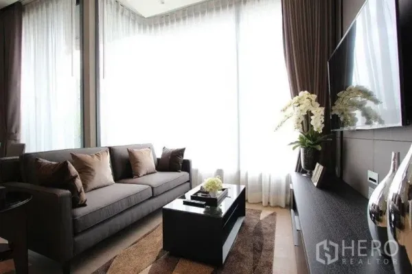 Condominium for rent in Watthana, Bangkok - Bright living room with grey sofa, coffee table and wall-mounted TV beside floor-to-ceiling curtains.