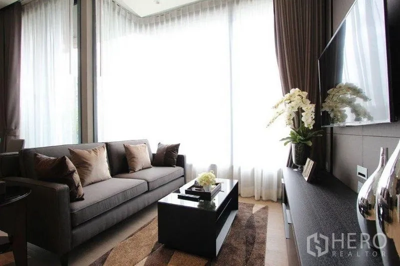 Condominium for rent in Watthana, Bangkok - Bright living room with grey sofa, coffee table and wall-mounted TV beside floor-to-ceiling curtains.