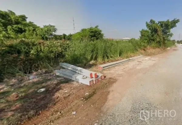 Land for sale in Khlong Toei, Bangkok - Roadside view of undeveloped plot with tall grasses and stacked concrete beams along a gravel lane.