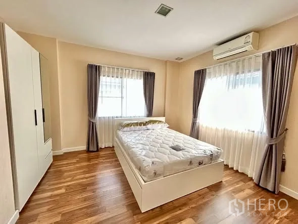Detached House for rent or sale in Prawet, Bangkok - Spacious bedroom with a double bed, wood floors, two windows, white wardrobe and air conditioner.